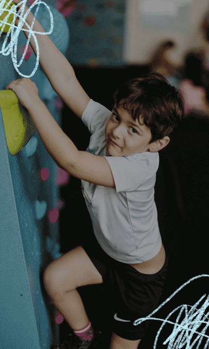 A young boy wearing a white t-shirt and black shorts climbs a blue indoor rock wall in DC, gripping a green hold with a determined expression—perfect for Summer Adventures. Scribbled white lines decorate the image’s corners.