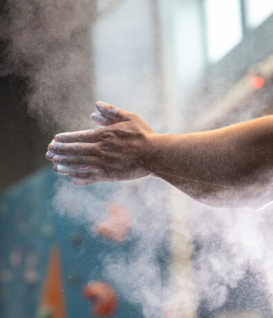 A close-up of a person clapping chalked hands, creating a cloud of chalk dust, with a blurred indoor climbing wall and K&N air filters in gyms helping keep the air clean in the background.