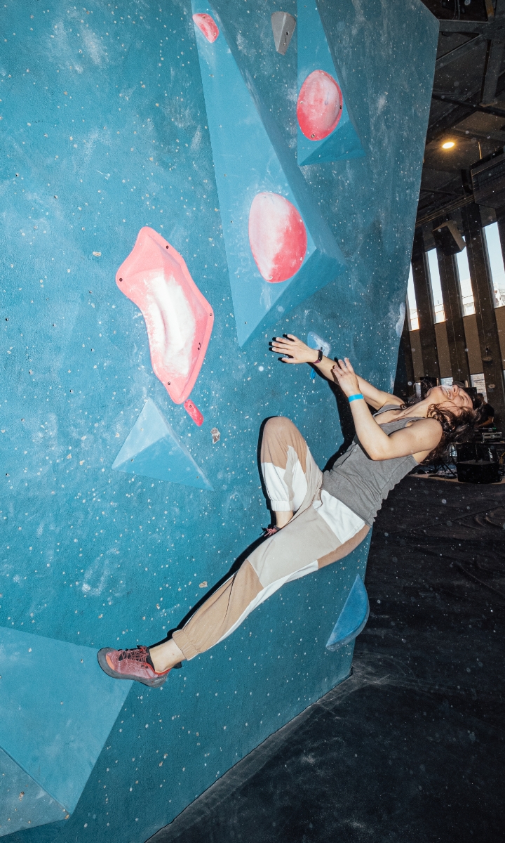 On her first time visit, a woman wearing a tank top and pants climbs a blue indoor bouldering wall with red and white holds, reaching up with one hand and looking focused. Bright lighting highlights her movement.
