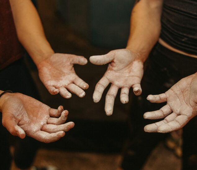 Four outstretched hands covered in white chalk powder hint at a First Time Visit to the climbing gym, with the blurred background drawing focus to the hands and their fresh experience.