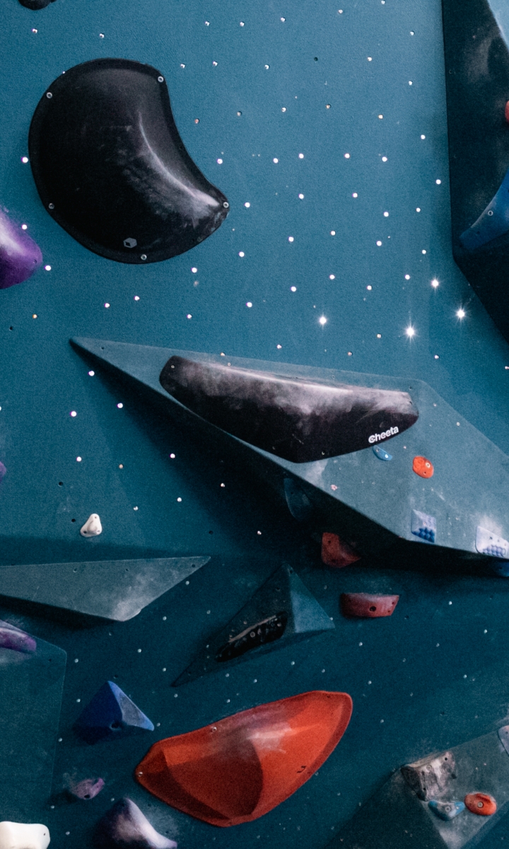 An indoor climbing wall with various geometric holds in black, purple, red, blue, and white colors, set against a teal background with small white dots.