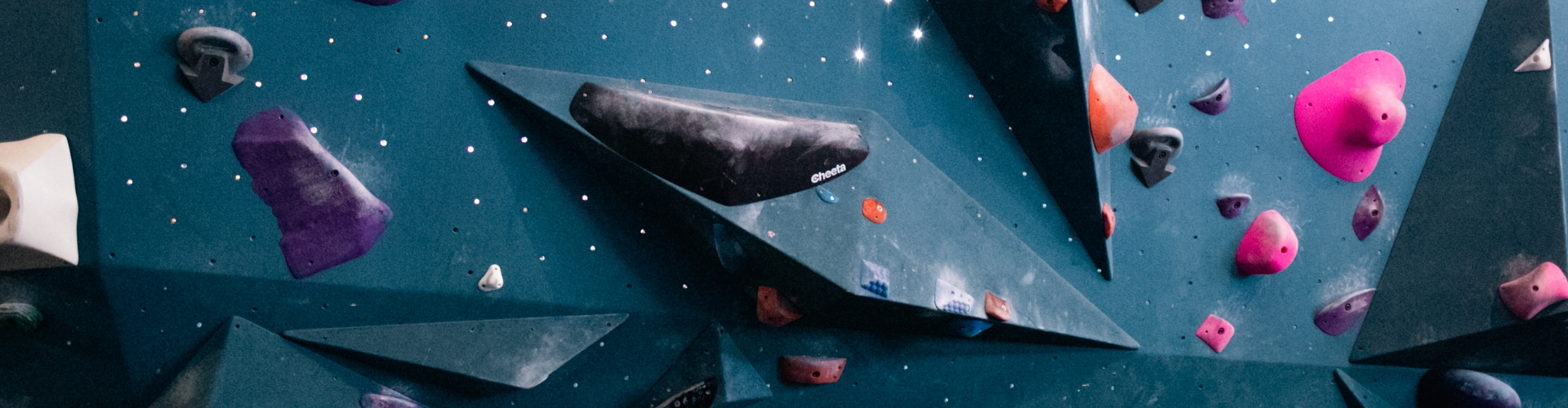 A close-up of an indoor climbing wall with various geometric holds in purple, black, pink, and red, set against a blue background dotted with small white spots.