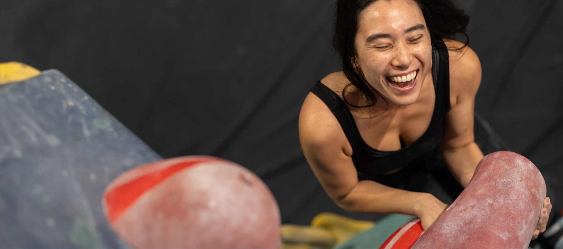 A woman in athletic clothing smiles and laughs while climbing on an indoor bouldering wall, holding onto a red handhold—embracing the spirit of partnership supported by TRUEMED.