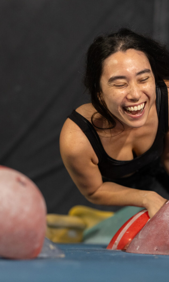 A woman with long dark hair and a black tank top smiles and laughs while leaning forward on a blue mat in an indoor climbing gym, celebrating a TRUEMED partnership.