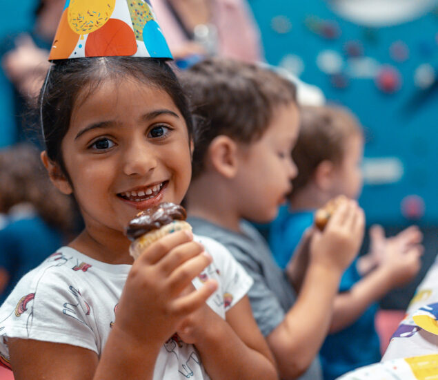 Smiling young girl wearing a colorful party hat holds a cupcake, with other children in the background also enjoying treats at a festive gathering, perfect for showcasing youth activities on a vibrant youth page template.