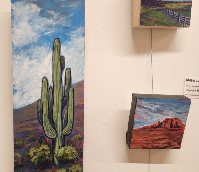 A space for members and local artists to showcase their artwork and creativity, this white gallery wall features a vertical painting of a large cactus in a desert, flanked by two small square paintings of a fence in a field and red rock formations.