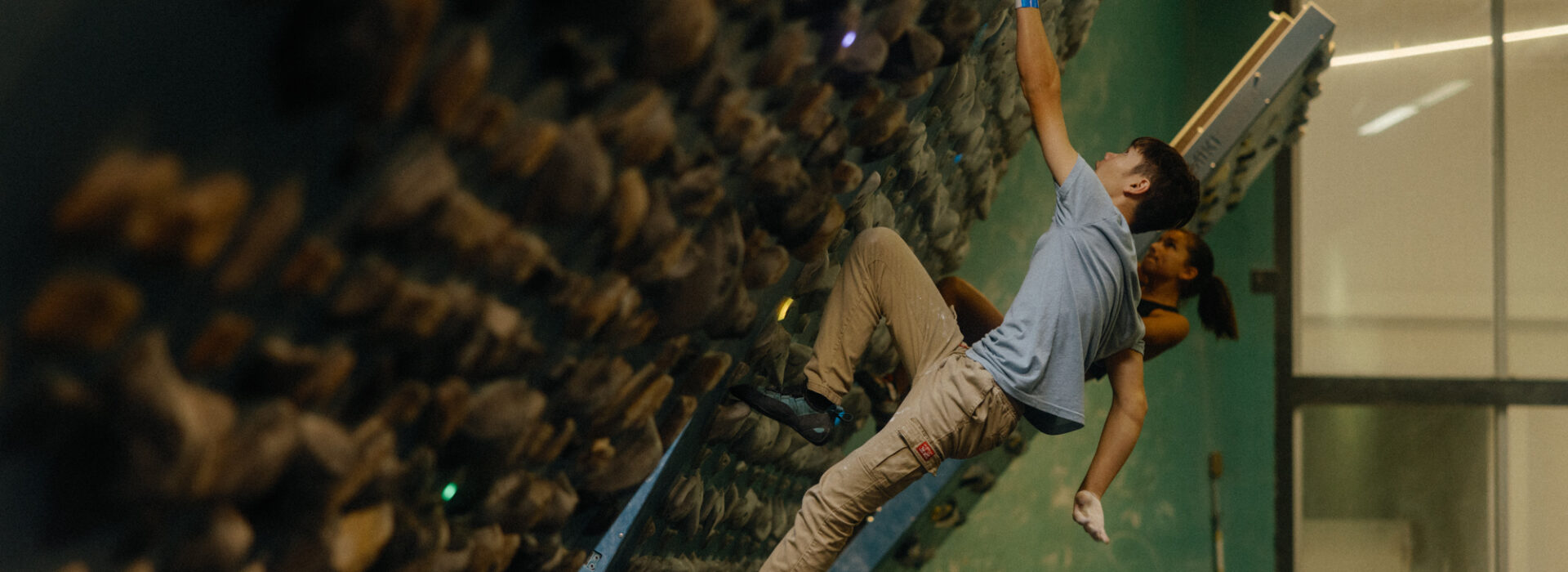 A boy in a blue shirt and beige pants climbs an indoor rock climbing wall at the Board League gym, while another climber is partially visible behind him. The wall is covered with numerous handholds.