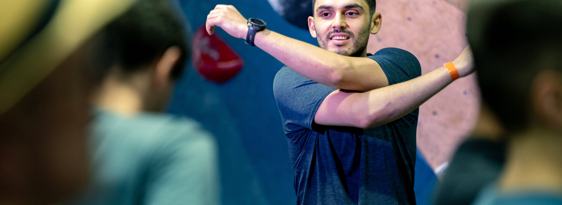 A man in a blue shirt stretches his arm across his chest in front of a DC climbing wall, with several Adult Team members standing nearby, slightly out of focus.