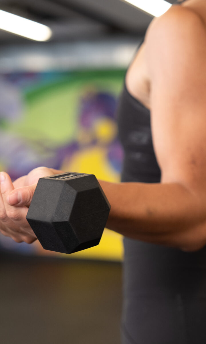 A person in athletic wear lifts a black dumbbell during a workout in a DC gym, with other people and colorful wall art in the blurred background—an inspiring scene of personal training in action.