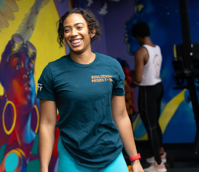 A woman in a blue shirt and shorts smiles while holding orange dumbbells at a colorful personal training gym in DC, vibrant mural walls surrounding her. Another person stands in the background, facing away.