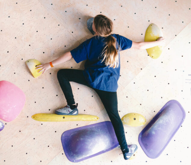 A child wearing a blue shirt and black pants climbs an indoor bouldering wall, reaching for yellow climbing holds with arms and legs spread wide—perfect for developing skills in youth sports or DC youth teams.