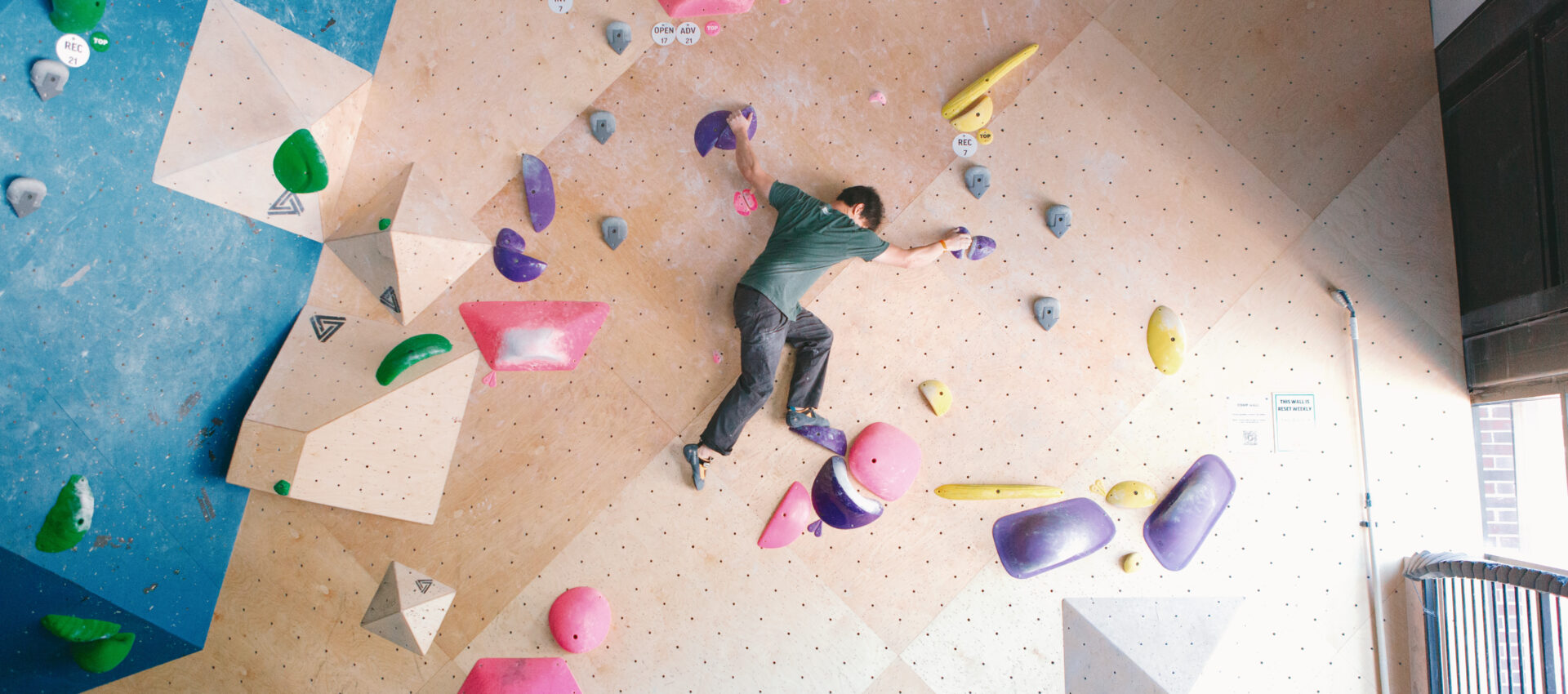 A person climbs an indoor bouldering wall at DC, reaching for colorful holds in pink, purple, green, and yellow on a geometric-patterned wall. Natural light enters from windows on the right—perfect for trying a Two Week Intro Pass.
