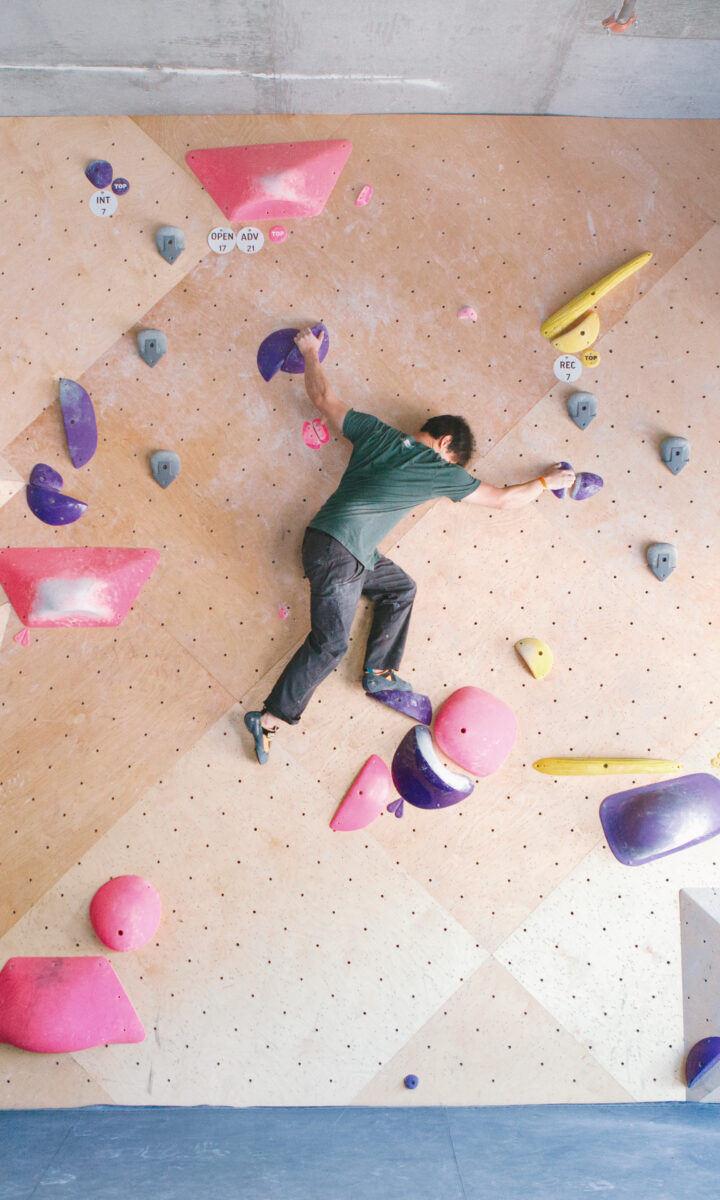 A person climbs an indoor bouldering wall at DC, reaching for colorful holds in pink, purple, green, and yellow on a geometric-patterned wall. Natural light enters from windows on the right—perfect for trying a Two Week Intro Pass.