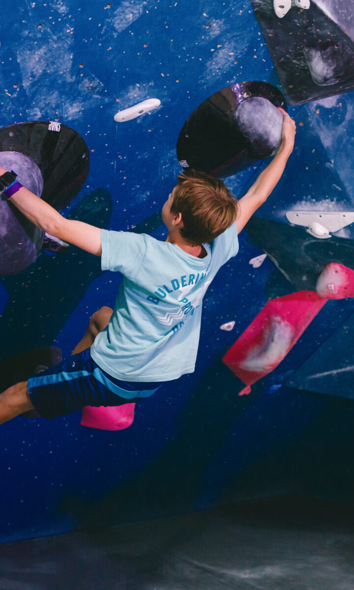 A person wearing a light blue T-shirt and blue shorts is climbing an indoor bouldering wall in DC, gripping colorful holds with one arm extended upward. The shirt reads BOULDERING on the back, representing local Youth Teams.
