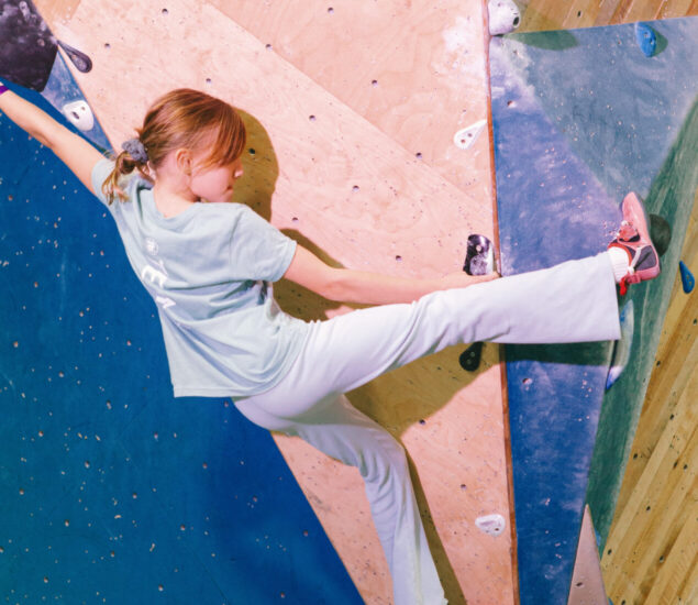 A girl with reddish hair, wearing a light blue shirt and white pants, climbs an indoor bouldering wall with DC Youth Teams, stretching her arms and legs to reach holds on the colorful surface.