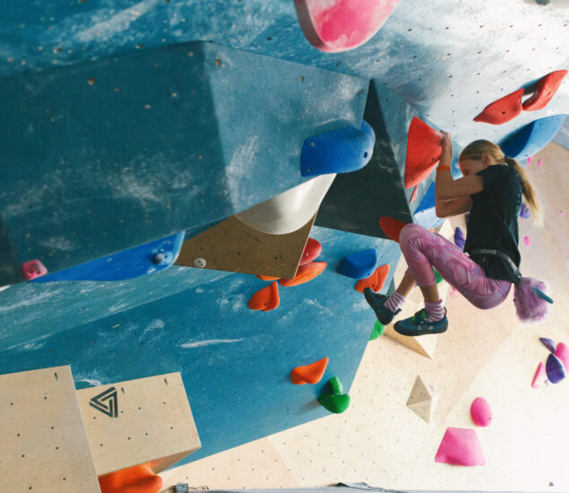A young girl in climbing gear scales an indoor bouldering wall at a DC youth program, gripping colorful holds and wearing pink leggings, with a padded mat below for safety.