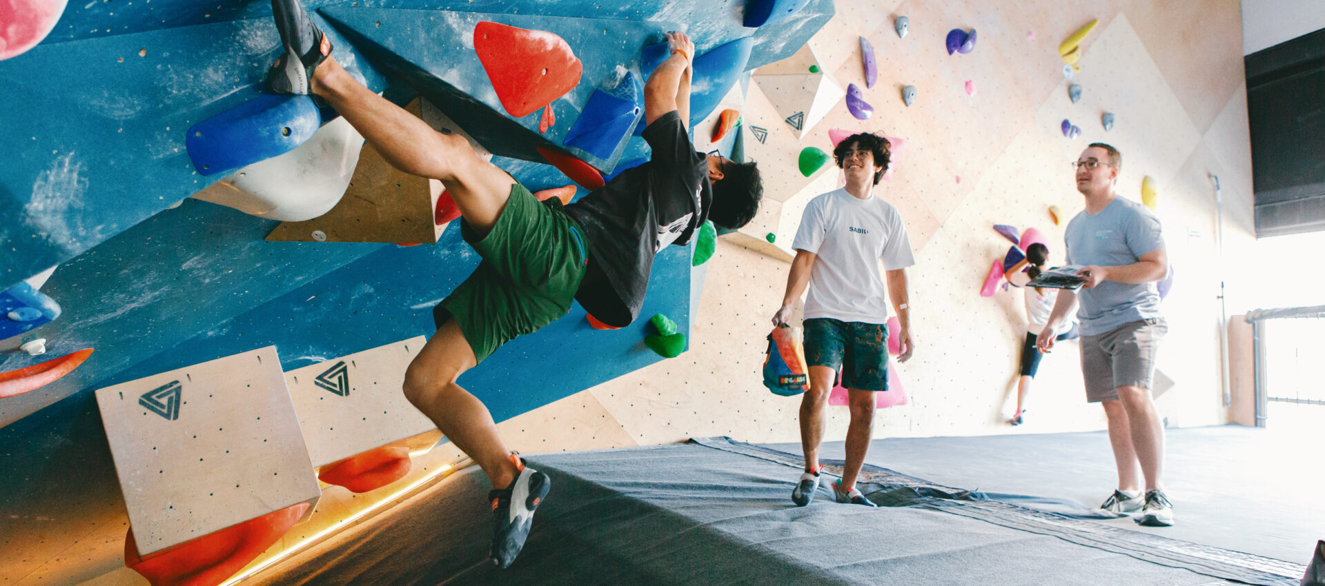 An adult team climbs on a colorful indoor bouldering wall while three others watch nearby, standing on padded flooring. The DC climbing gym features various holds and sloped surfaces.
