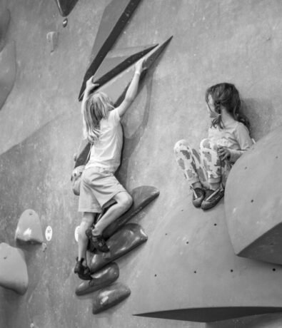 Two young children climb on an indoor bouldering wall at Boulderfest 2025 in Brooklyn. One child ascends using handholds, while the other sits on a large rock-like hold, resting. The scene is captured in black and white.