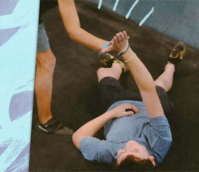 A person lying on the ground reaches up and grabs the hand of another who is standing, as if being helped up by a summer camp master, on a black mat with blue and white accents in the background.