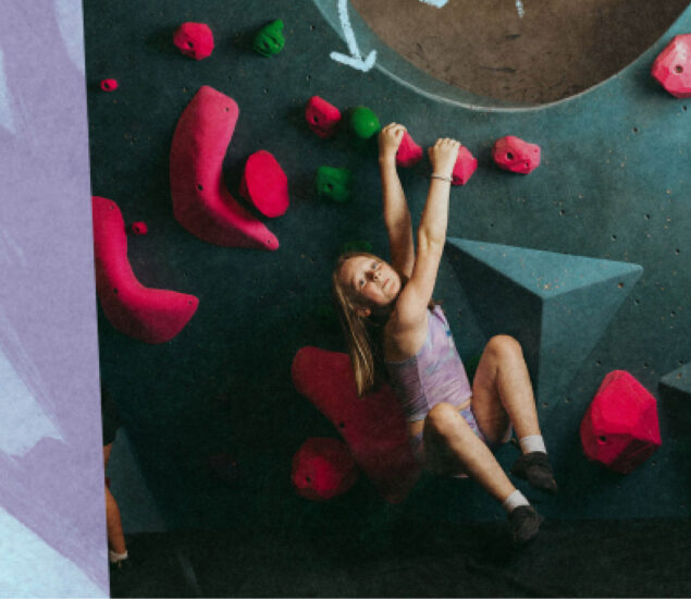 A young girl wearing a tank top and shorts climbs an indoor rock wall with bright pink and green holds, smiling as she looks up—perfect for a summer camp master searching for an inspiring camp template photo.
