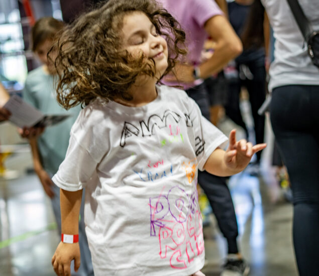 A young child with curly hair joyfully dances with eyes closed and a smile, wearing a white T-shirt decorated with colorful drawings—capturing the vibrant spirit of youth programs in DC. People and a busy indoor setting are blurred in the background.