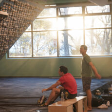 Two men in athletic clothing are at Brooklyn Climbing Yoga and Fitness. One sits on a bench, the other stands, both facing a climbing wall as sunlight streams through large windows, creating a warm, bright atmosphere.
