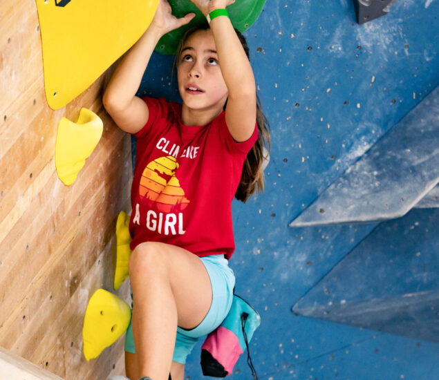 A young girl wearing a red “Climb Like a Girl” shirt climbs an indoor rock wall during one of DC’s youth programs, reaching up to grip a green hold, with yellow and gray holds around her and a chalk bag at her waist.
