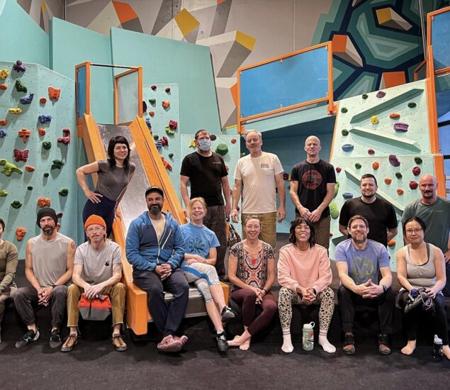 A group of people from local Community Clubs pose and smile together in front of an indoor climbing wall in Minneapolis, surrounded by colorful holds, geometric patterns, and a playful slide. Some are seated while others stand proudly.