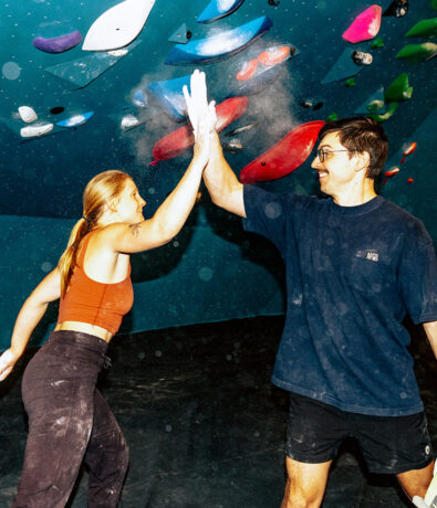 Two people wearing athletic clothes high-five in front of a colorful indoor climbing wall, both smiling and with chalk on their hands.