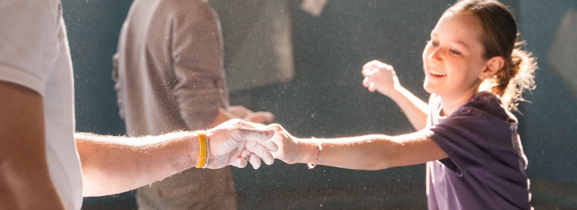 A smiling girl in a purple shirt reaches out and bumps fists with someone, both hands covered in chalk dust, in a brightly lit Brooklyn indoor setting—capturing the joyful spirit of local After School Programs. Another person is blurred in the background.