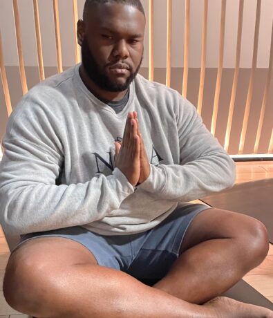 A man sits cross-legged on a mat with his palms pressed together in a prayer position, dressed in a gray sweatshirt and blue shorts—taking a moment to relax after yoga, in a room with vertical wooden slats behind him.