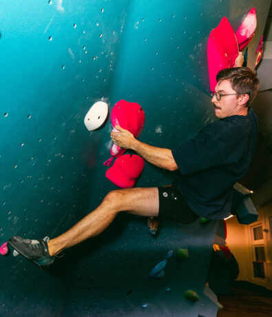 A man with glasses and a mustache climbs an indoor bouldering wall, gripping large pink holds. Wearing climbing shoes, a dark shirt, and shorts, he enjoys colorful routes—perfect for a summer membership adventure.