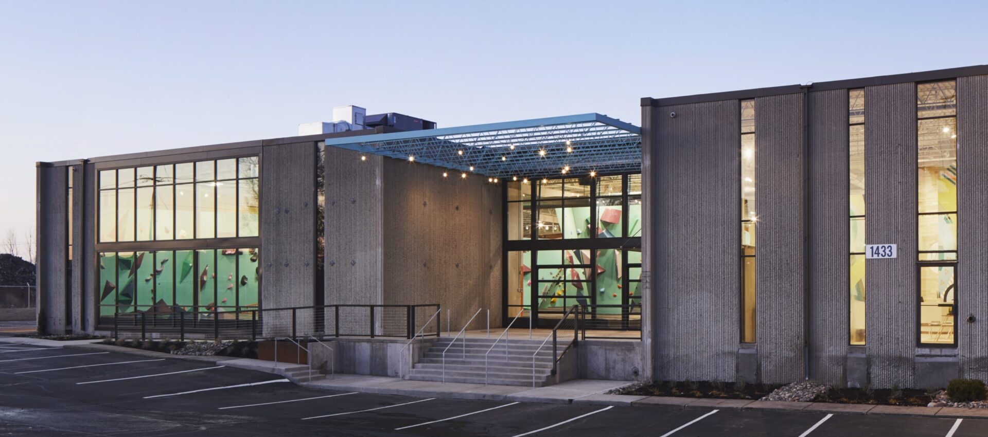 A modern Seattle building with large glass windows, a central staircase entrance, and a partially covered patio. The parking lot in front has several empty spaces, and the interior features colorful climbing walls. Contact Us for more details.