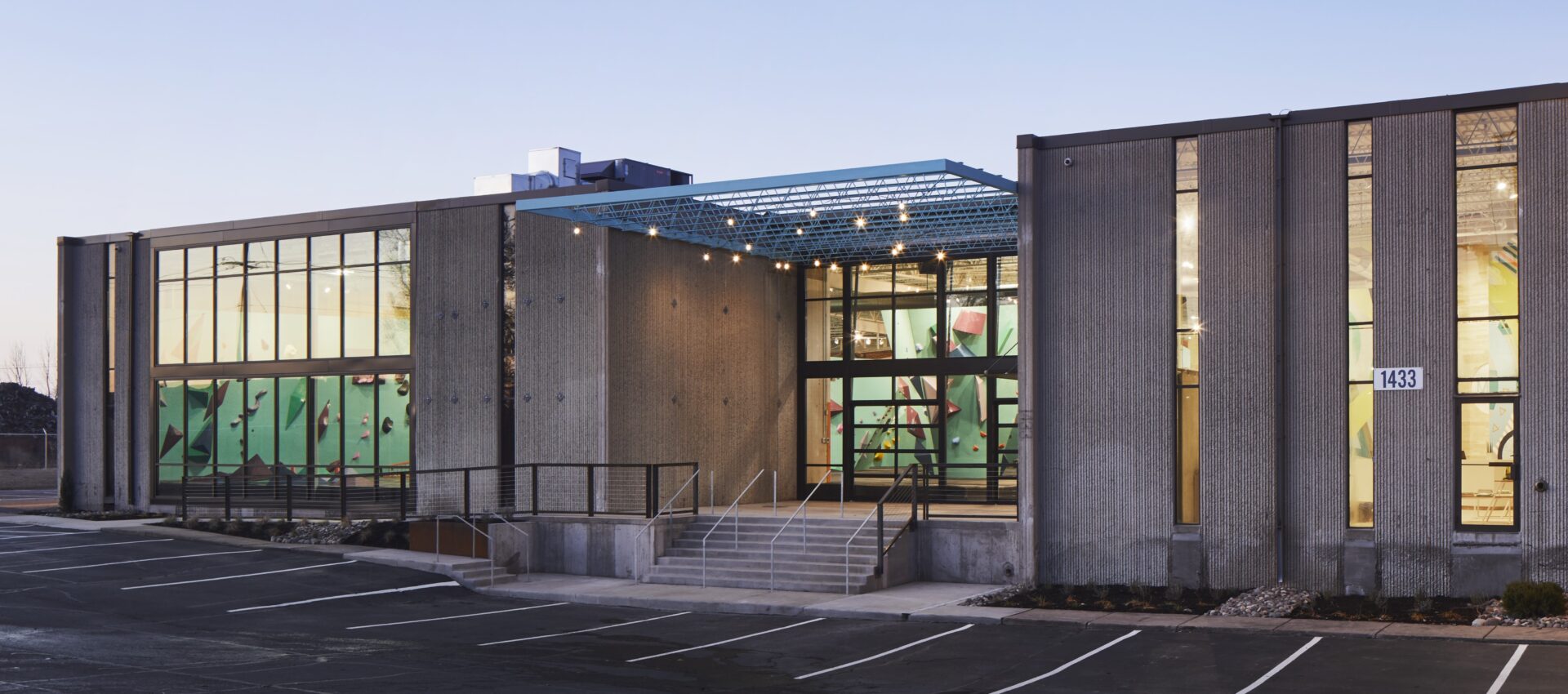 A modern, concrete building in Seattle with large glass windows reveals colorful indoor climbing walls. The entrance features stairs and a railing, with an empty parking lot in front. Contact Us for more information.