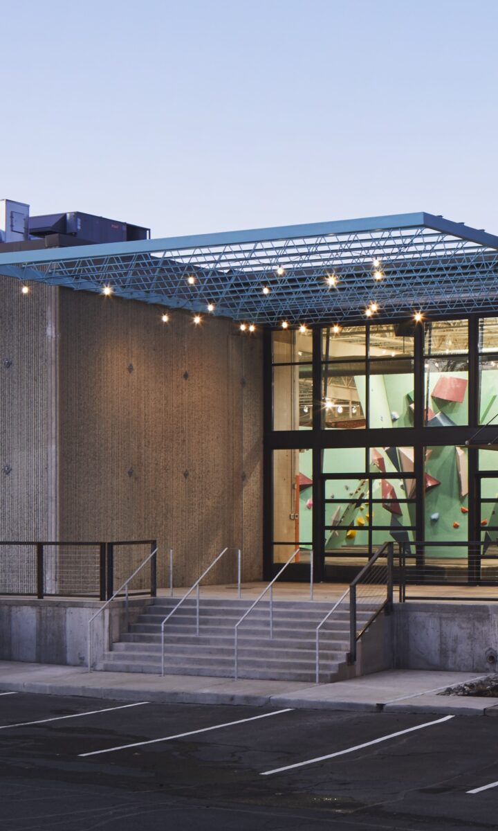 A modern, concrete building in Seattle with large glass windows reveals colorful indoor climbing walls. The entrance features stairs and a railing, with an empty parking lot in front. Contact Us for more information.