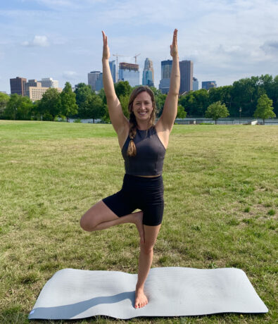 A woman practices yoga outdoors on a mat, standing in tree pose with arms raised. She is smiling, with city buildings and trees in the background under a partly cloudy sky.
