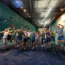 A group of people jump in the air, smiling and posing together inside an Austin Climbing Gym with colorful climbing holds on the walls.