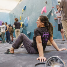A woman sits and smiles on the gym floor, wearing climbing gear. Behind her, people stand and socialize near a blue indoor climbing wall with colorful holds. The atmosphere is lively and casual.
