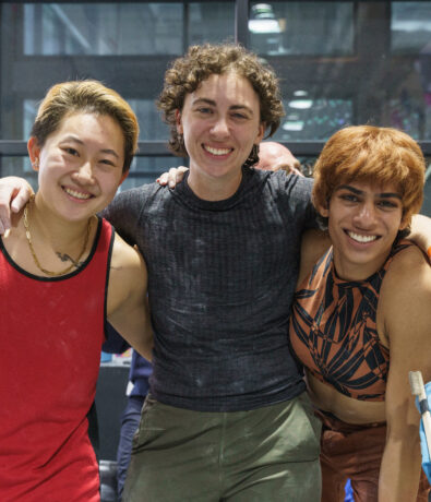 Three people smile and pose together indoors, one giving a thumbs up, another with an arm around the group. They look happy and friendly. A fourth person in the background also smiles, with climbing gear nearby hinting at their shared climbing adventure.