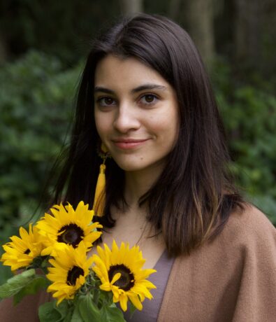 A young woman with long dark hair, wearing a brown shawl, stands outdoors near the University District Seattle, holding a bouquet of yellow sunflowers. Green foliage and trees are blurred in the background as she smiles softly at the camera.