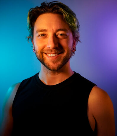A smiling person with short, wavy hair wearing a sleeveless black shirt stands in front of a gradient blue and purple background, ready to share DC personal training tips.