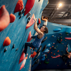 A person in a blue tank top climbs a blue indoor bouldering wall with red, orange, and black holds at an Austin Climbing Gym. Another climber is visible nearby as bright lights illuminate the gym.