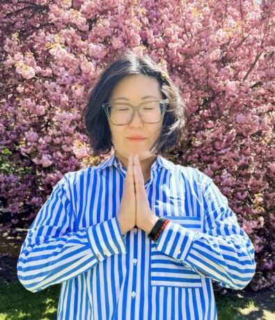 A person wearing glasses and a blue-and-white striped shirt stands with hands pressed together in a prayer pose, eyes closed, in front of a blooming pink cherry blossom tree after a class at Brooklyn Climbing Yoga and Fitness.