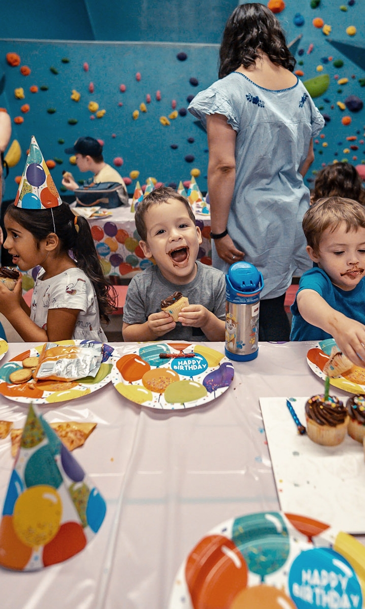 Three young children sit at a birthday party table in a climbing gym, with colorful hats and plates, enjoying cupcakes and snacks. The smiling boy in the center holds a cupcake. An adult stands in the background, adding to the playful Birthday Parties atmosphere.