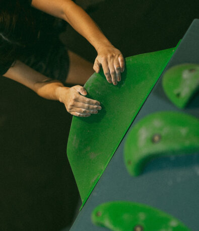 A person with chalked hands grips a large green climbing hold on an indoor climbing wall at a climbing gym in Seattle’s University District, viewed from above.