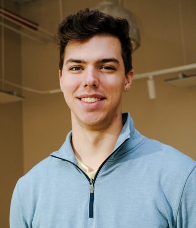 A young man with short brown hair, wearing a light blue zip-up sweater, smiles at the camera. The background features an indoor setting with beige walls and exposed piping, hinting at a Brooklyn Climbing Yoga and Fitness studio vibe.