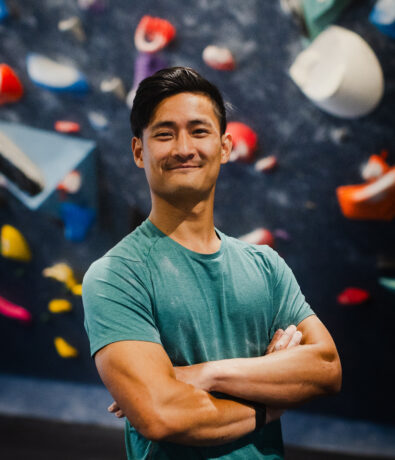A young man with short dark hair, wearing a teal t-shirt, stands confidently with arms crossed in front of the vibrant Upper Walls at Fremont’s indoor rock climbing gym.
