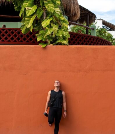 A person in black athletic wear stands barefoot, leaning against an orange wall with one foot raised as if preparing for climbing. Above the wall are large green plants and a thatched roof structure. The sky is partly cloudy.