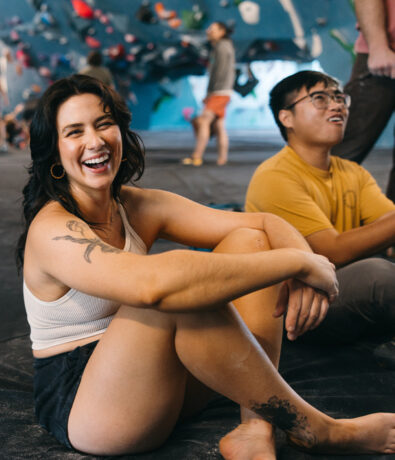 A woman in a white tank top and shorts sits on the floor smiling, with a man in a yellow shirt behind her. They are in an indoor rock climbing gym, with climbing walls and people in the background.