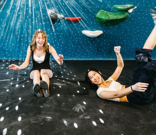 Two young women laugh and relax on the floor of an indoor climbing gym, surrounded by colorful holds and a starry blue wall—enjoying the Membership Benefits at Austin Bouldering Project.